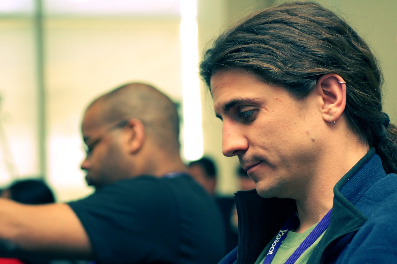 A person with long hair and a lanyard looks down thoughtfully, while another person is blurred in the background.