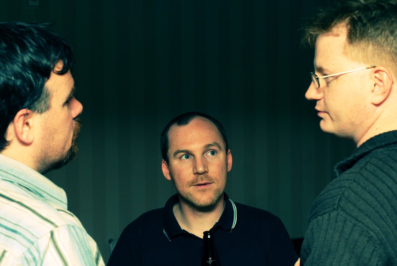 Three men engaged in conversation indoors, with one holding a bottle and looking attentively at the others.