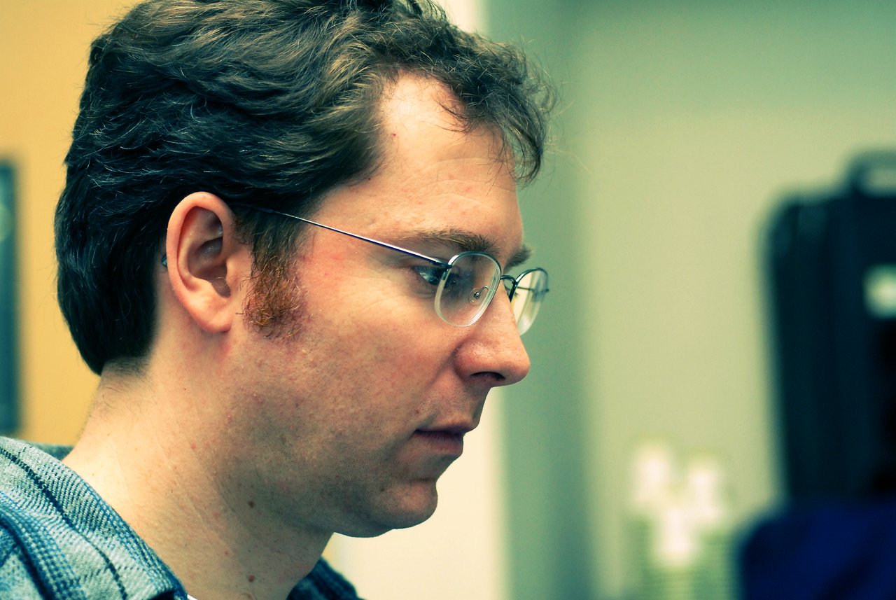 A man with glasses and short brown hair looks intently at something off-camera in an indoor setting.