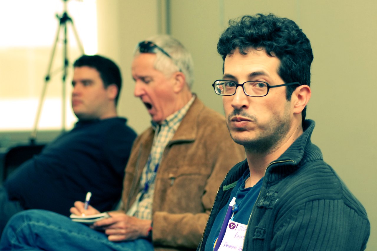 A man wearing glasses and a name tag looks at the camera, while others in the background take notes.