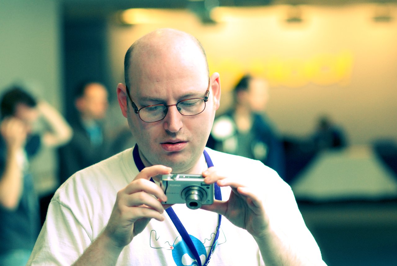 A person wearing glasses and a lanyard looks at a digital camera, possibly reviewing a photo.