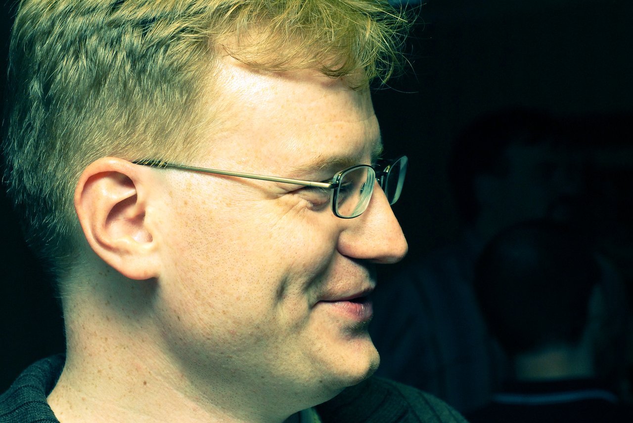 A man with glasses and short blond hair smiles while looking to the side in a dimly lit setting.