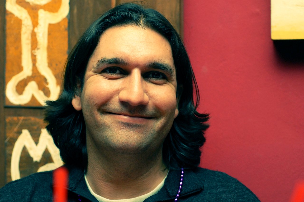 A person with shoulder-length dark hair smiles at the camera, wearing a dark sweater and a beaded necklace.