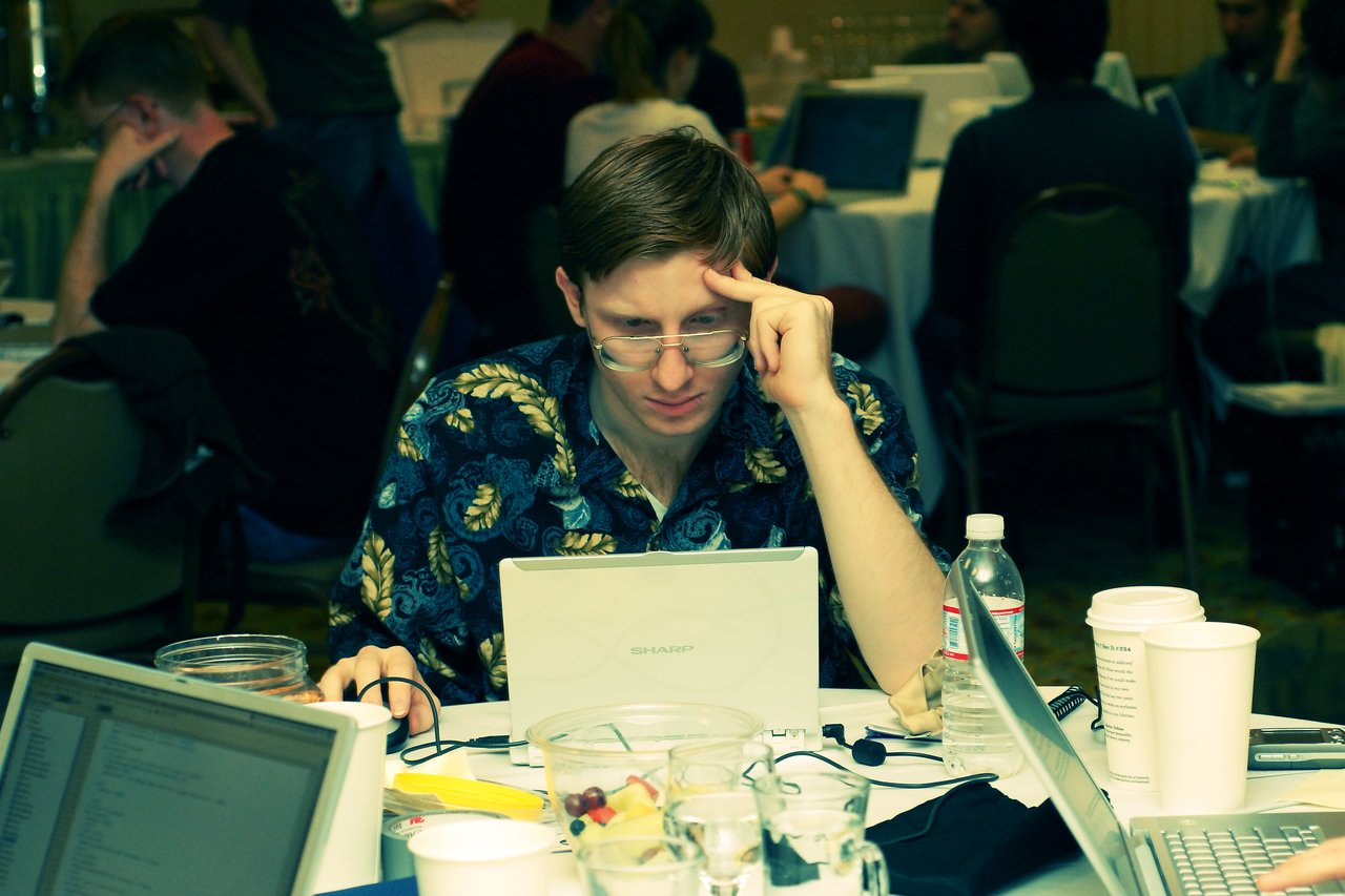 A man in a patterned shirt focuses intently on his laptop at a conference table with drinks and laptops.