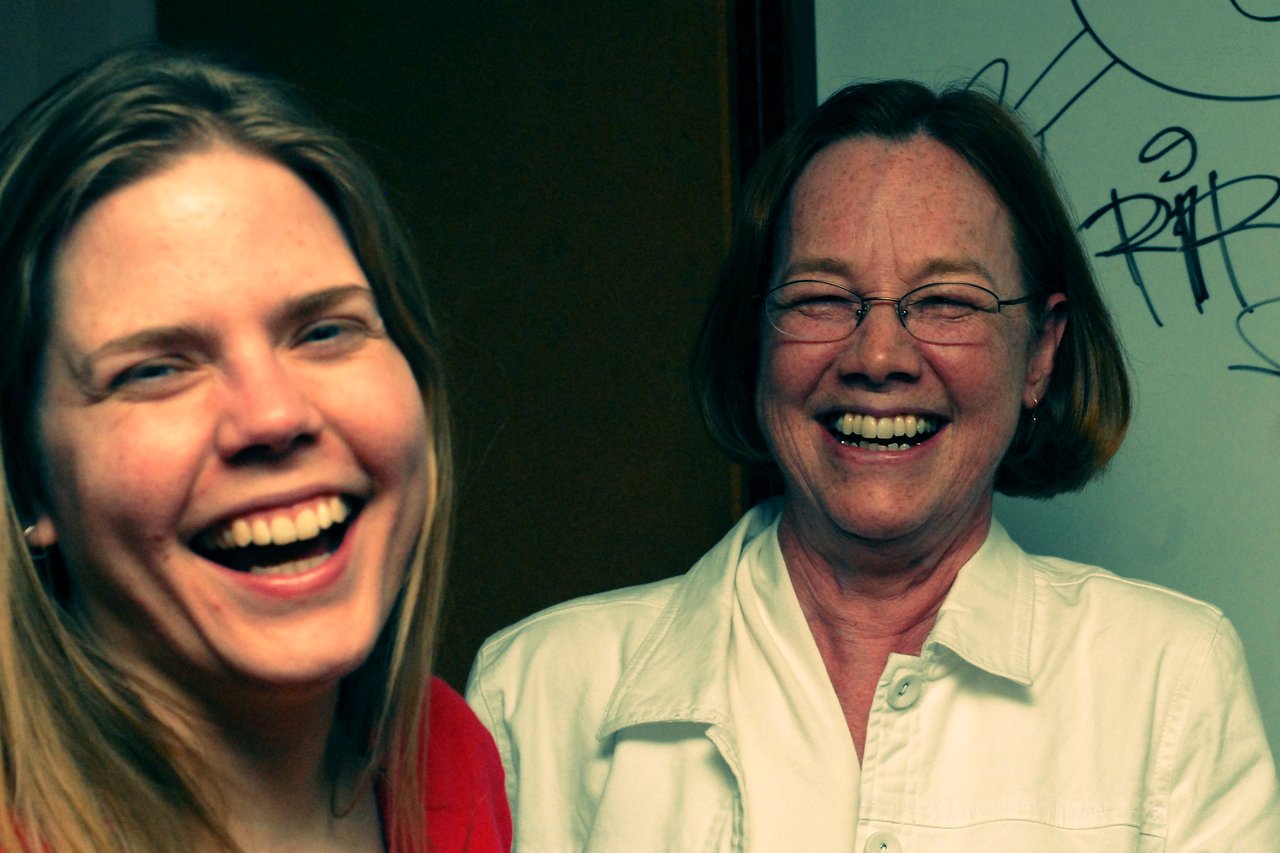 Two women laughing together, one in a red top and the other in a white jacket with glasses.