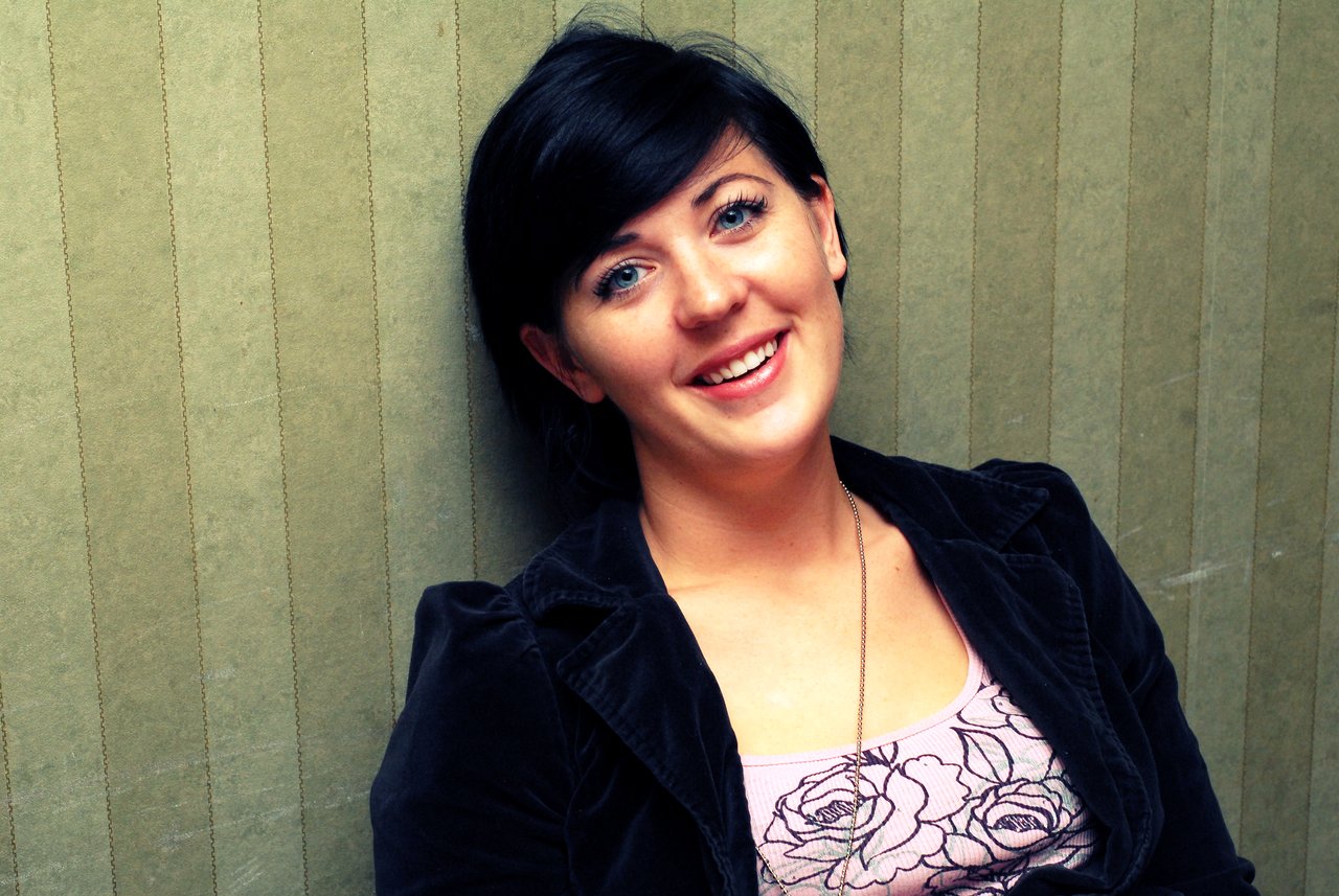 A person with short dark hair smiles while leaning against a textured green wall, wearing a black jacket and floral top.