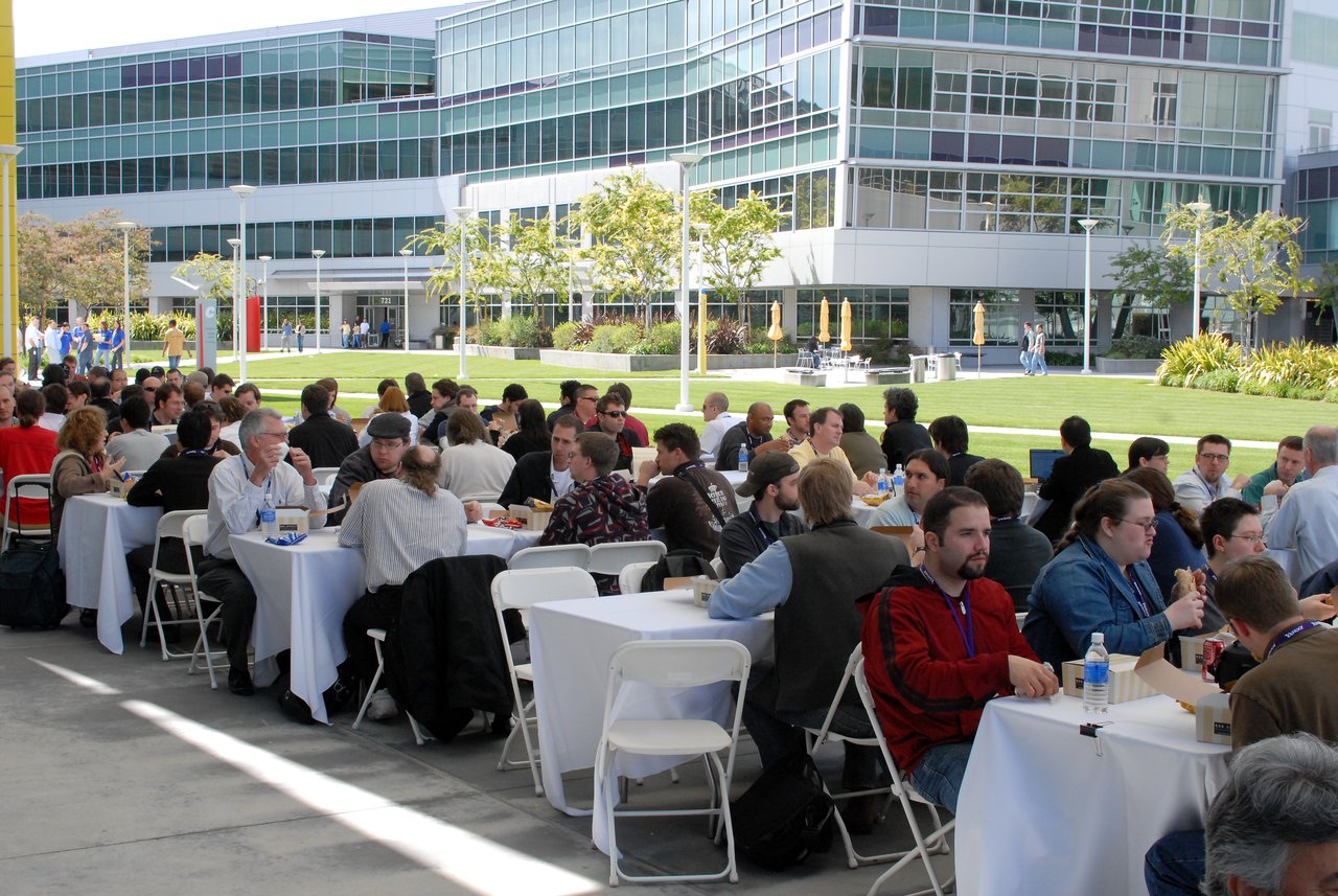 A large group of people sits at outdoor tables, eating lunch and talking during a conference event.