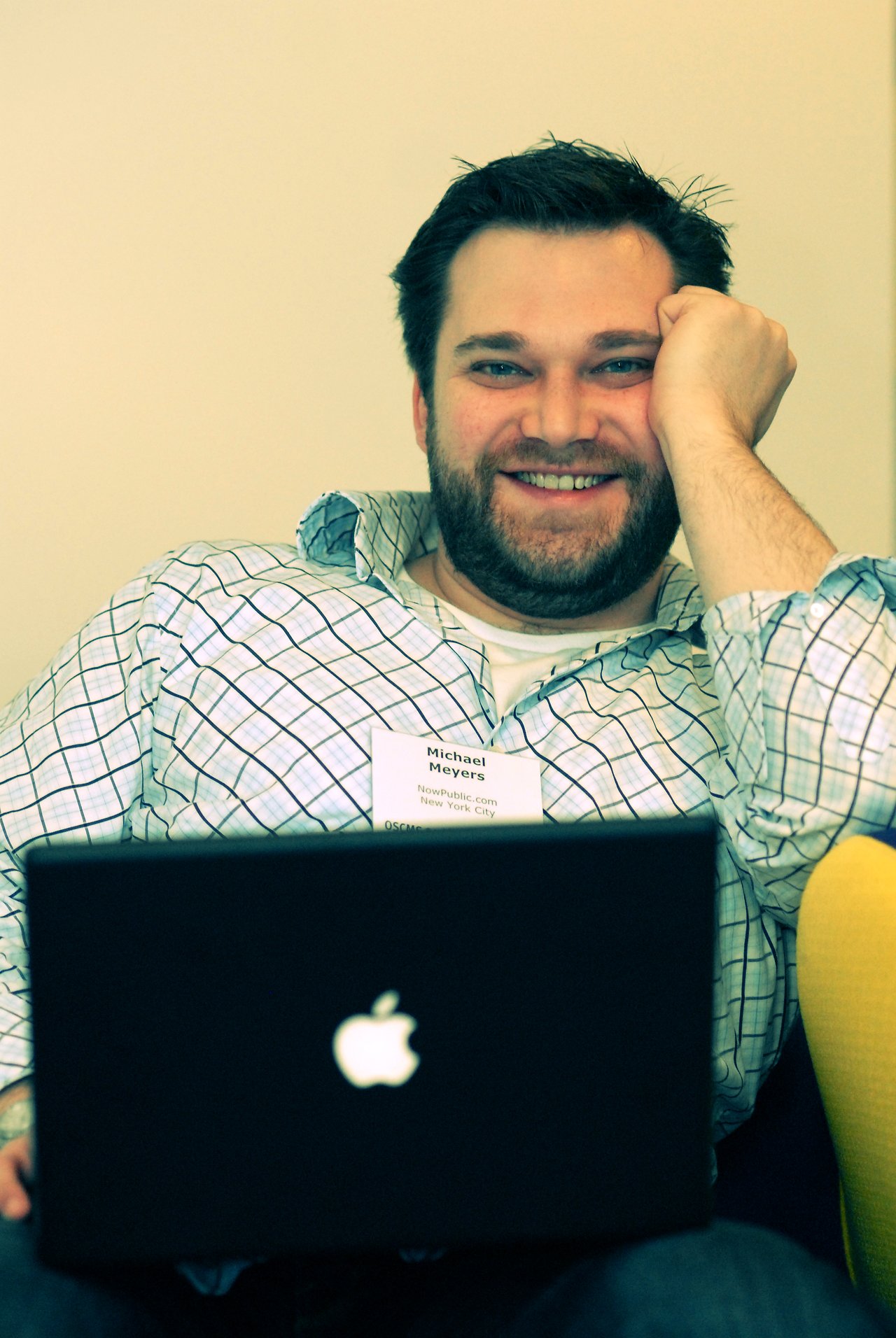 A man with a name tag smiles while sitting with a laptop on his lap at a conference.