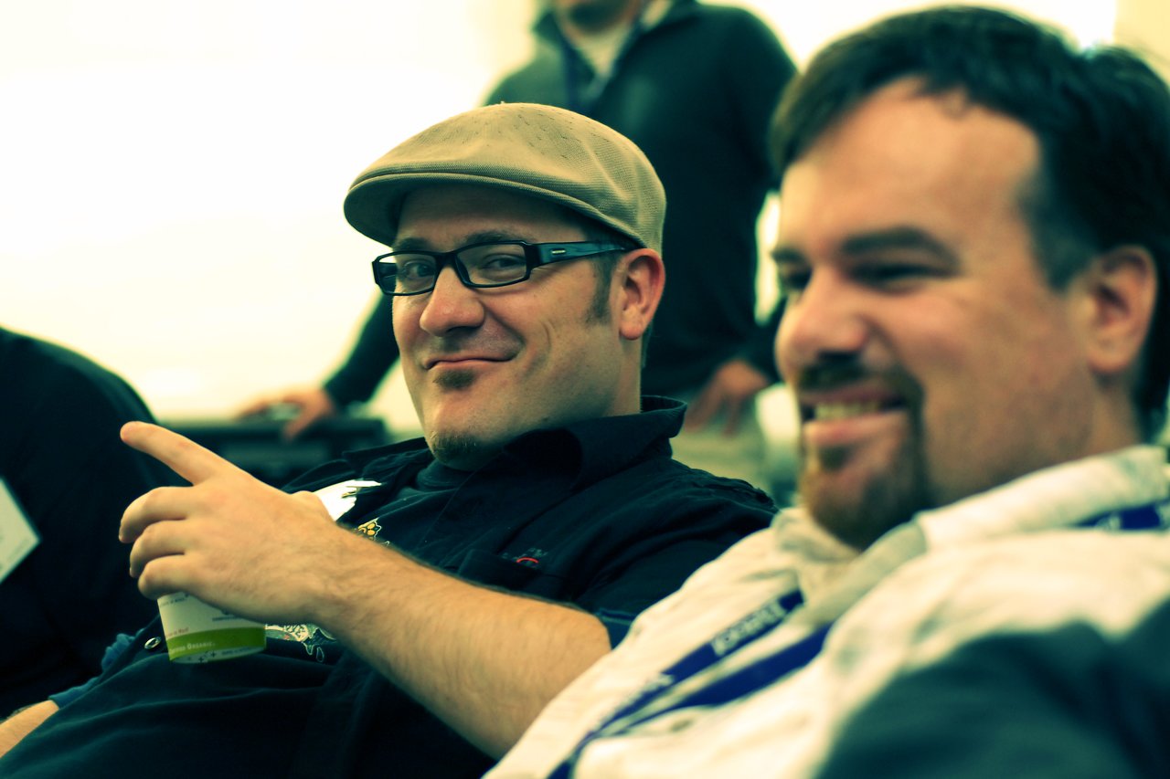 Two men sitting and smiling, one wearing a cap and glasses while pointing and holding a drink.
