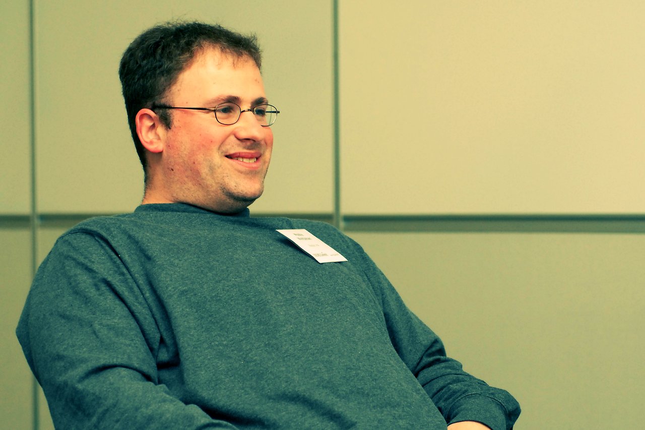 A man wearing glasses and a name tag sits and smiles in a conference setting.