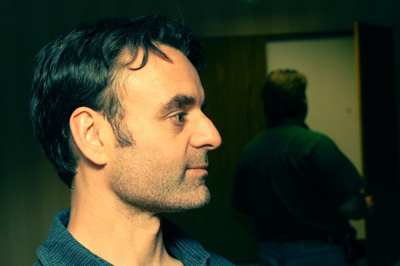 A man in profile looks to the left, while another person stands in the background facing away.