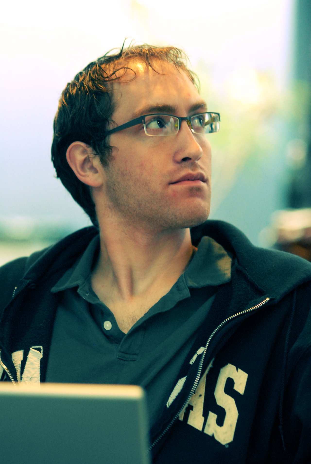 A person wearing glasses and a hoodie looks to the side while sitting in front of a laptop.