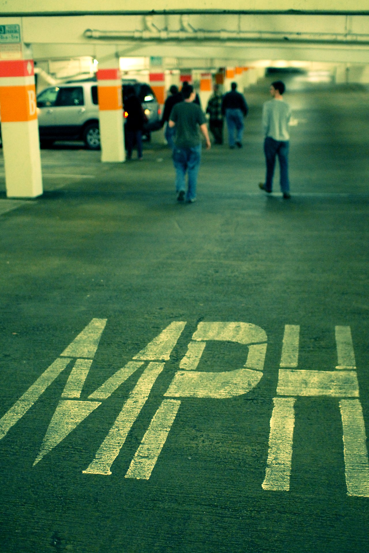 People walk through a dimly lit parking garage with "MPH" painted on the ground in the foreground.