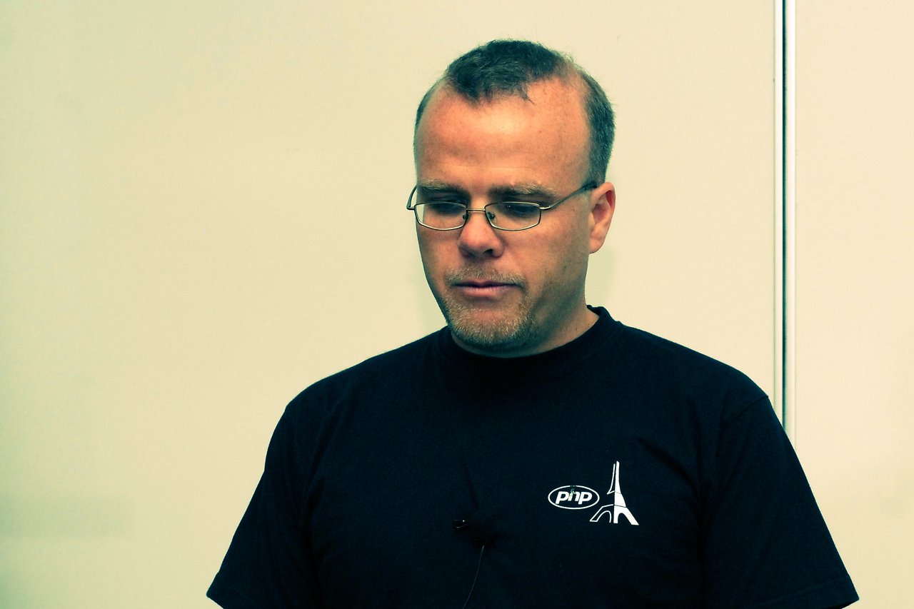 A man wearing a black PHP t-shirt and glasses looks down with a focused expression in a conference setting.