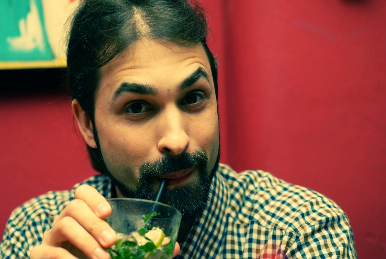 A person with a beard and checkered shirt sips a drink through a straw, looking directly at the camera.