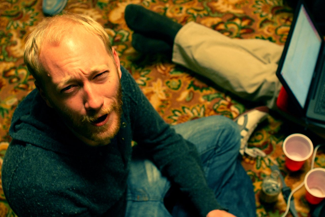 A man with a beard sits on the floor, looking up with a puzzled expression.