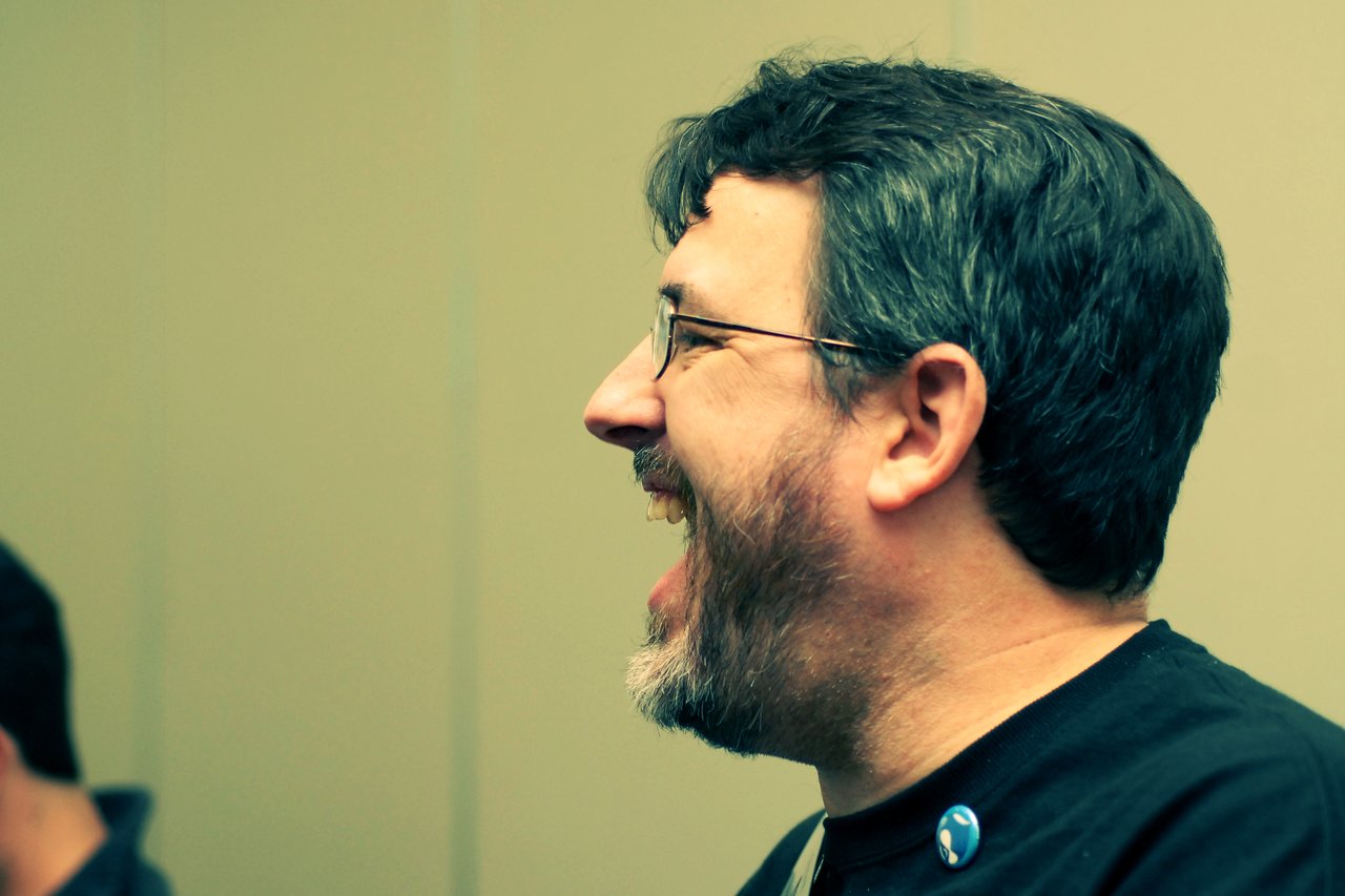 A man with glasses and a beard is laughing while wearing a black shirt with a Drupal pin.