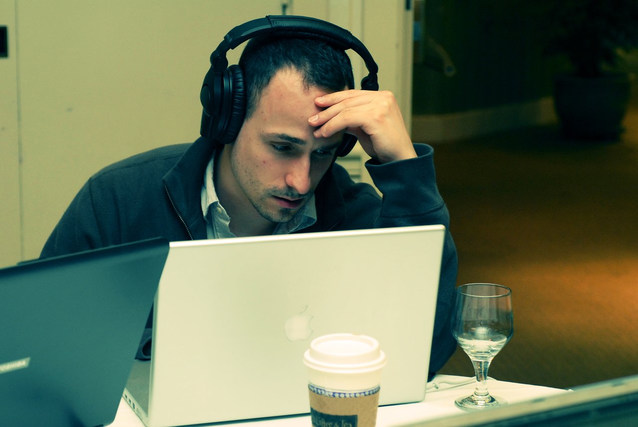 A person wearing headphones looks focused while working on a laptop, with a coffee cup and glass nearby.