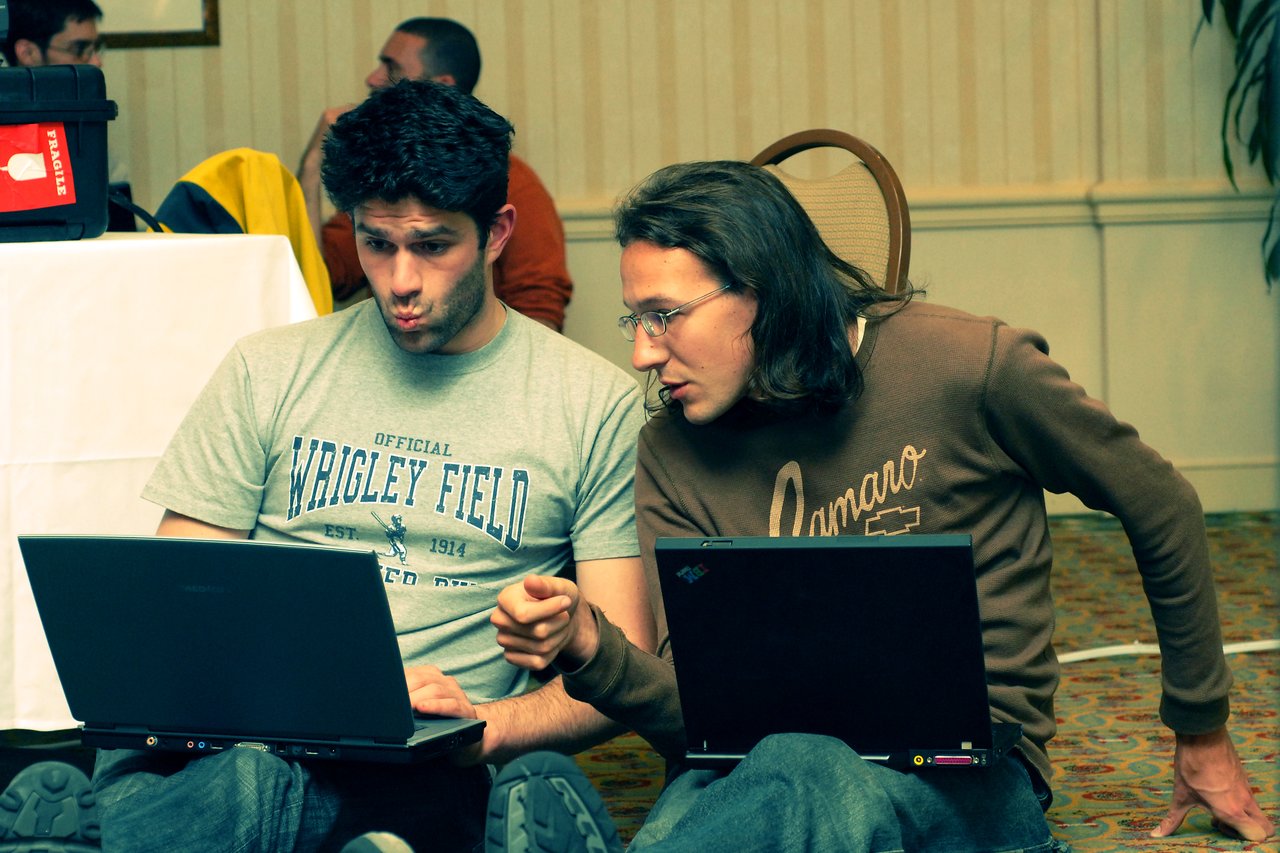 Two people sitting on the floor with laptops, discussing something on the screen while one gestures and the other listens.
