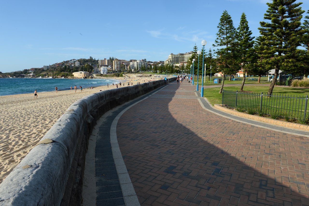 A paved walkway curves along the beach, with people walking, jogging, and relaxing on the sand near the ocean.