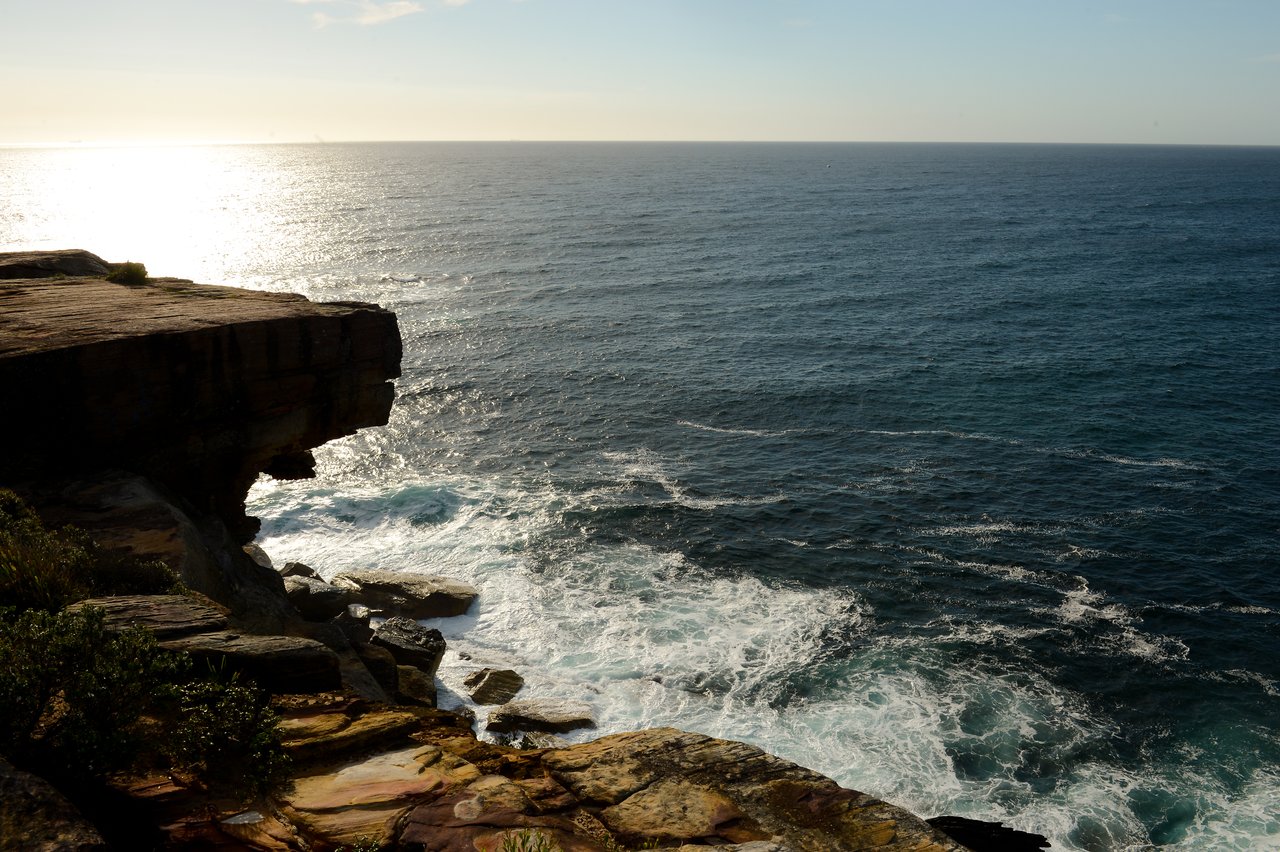 Rocky cliff overlooking the ocean with waves crashing against the shore under a bright sky.