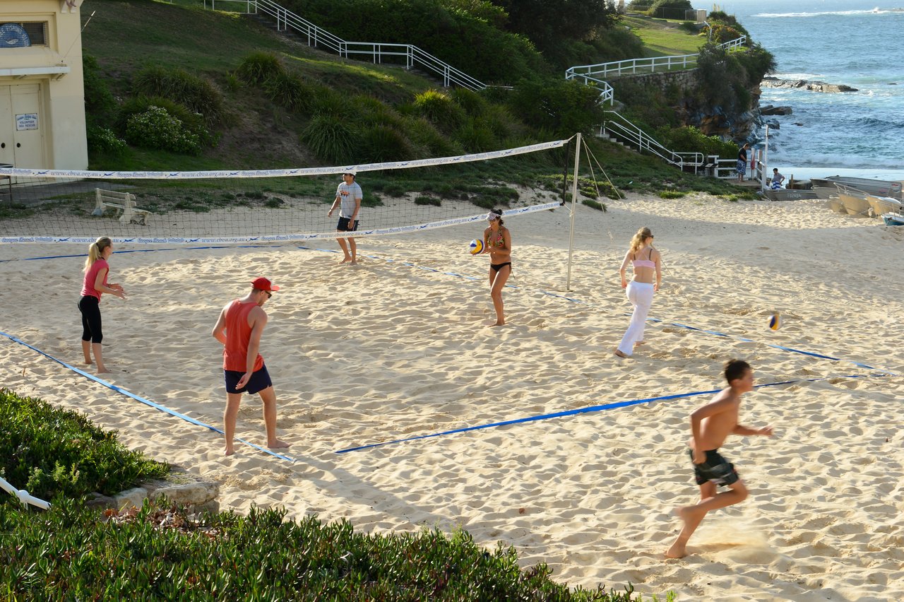 People playing beach volleyball on the sand near the ocean, with some actively hitting the ball and others watching.