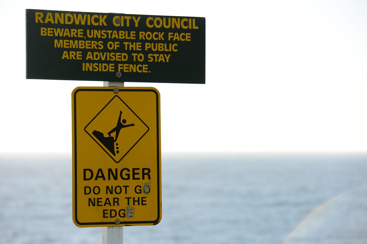 Warning sign near the ocean cautioning about unstable rocks and advising people to stay away from the edge.