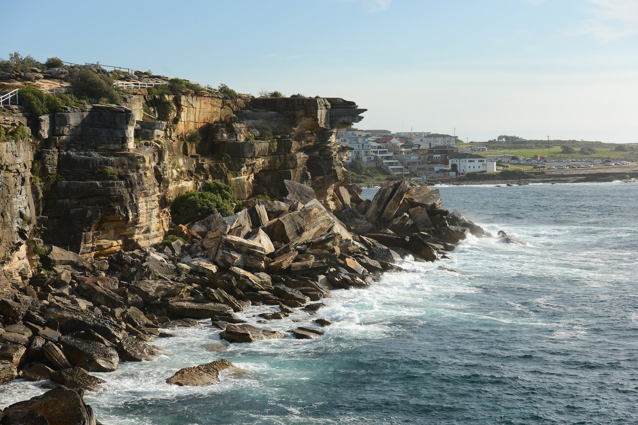 Rocky cliffs meet ocean waves at Coogee Beach, with houses and a parking area visible in the background.