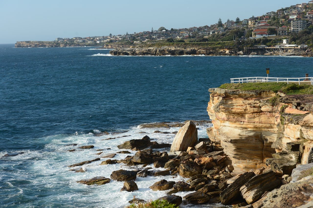 Rocky coastline with ocean waves crashing against cliffs, a white fence on the edge, and houses in the background.