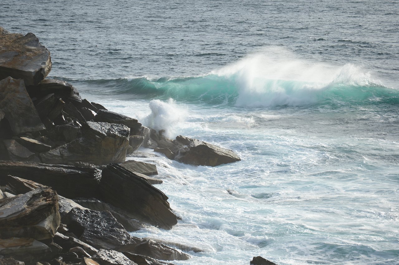 Waves crash against rocky cliffs at Coogee Beach, creating white foam and splashes in the ocean.