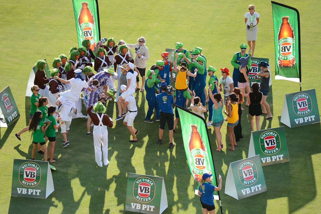 A group of people in costumes and cricket attire celebrate on a field, surrounded by Victoria Bitter banners.