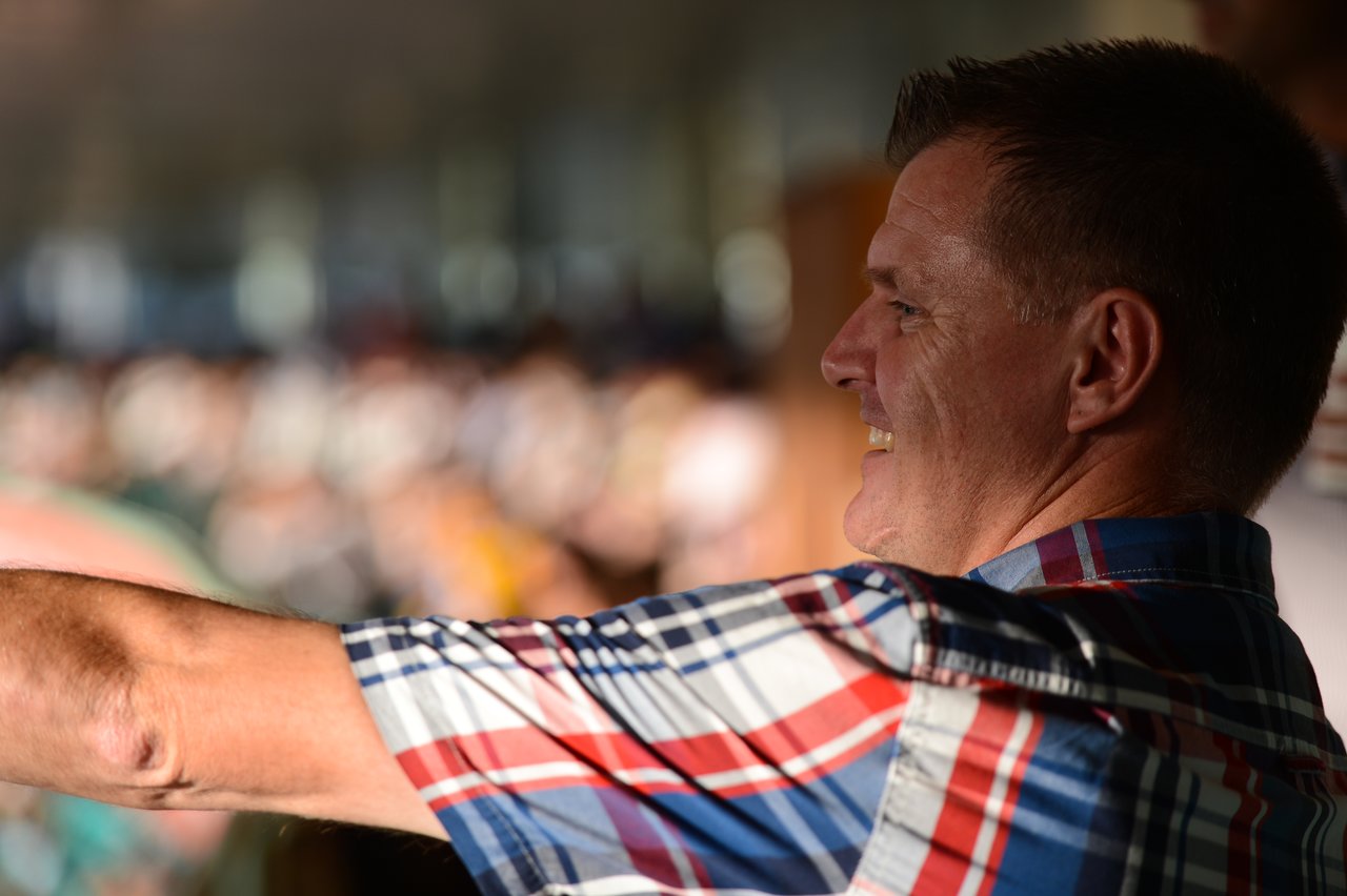 A man in a plaid shirt smiles and gestures while watching a cricket match in a crowded stadium.