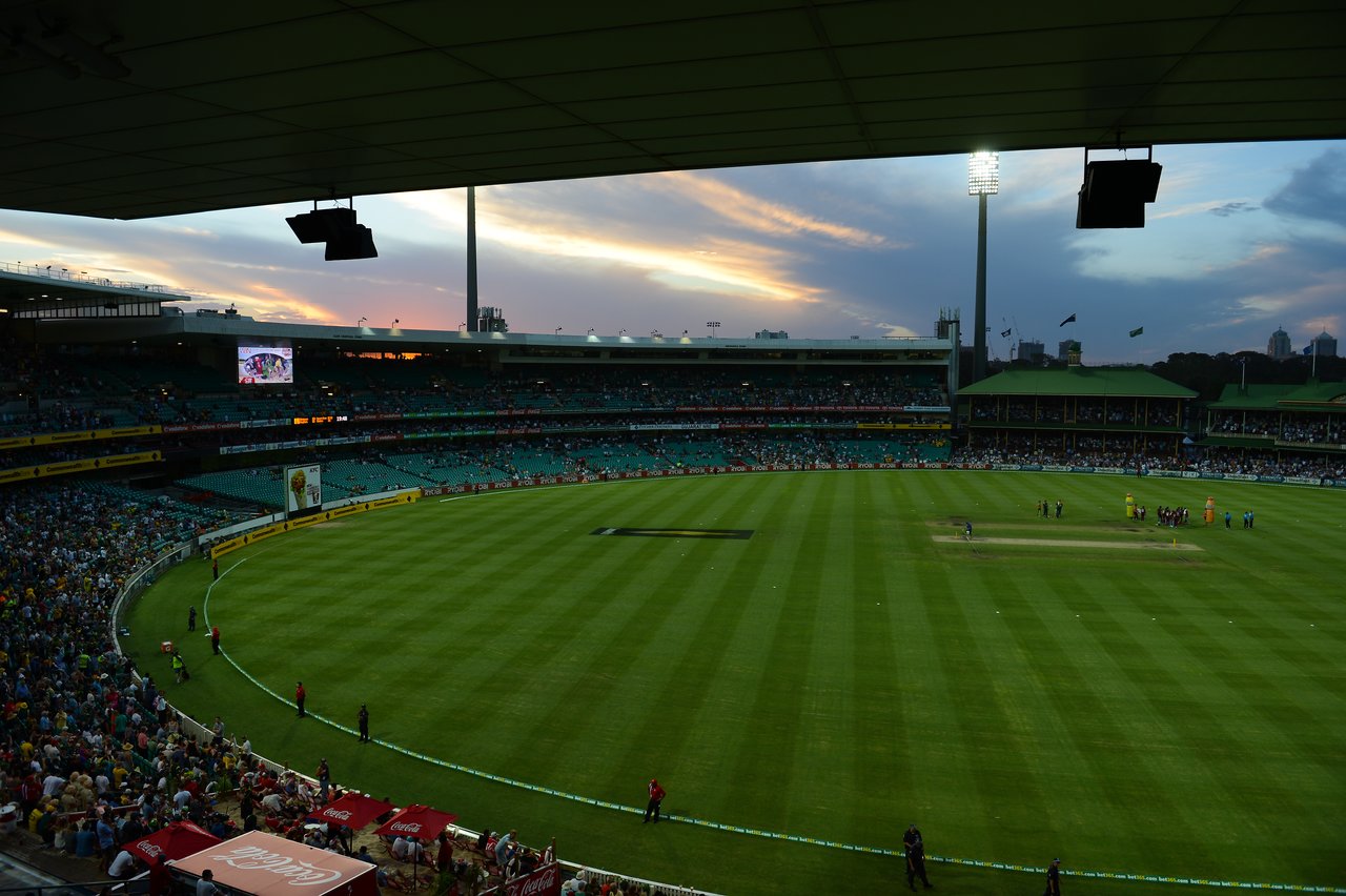 A cricket match at a stadium with players on the field and spectators in the stands during sunset.