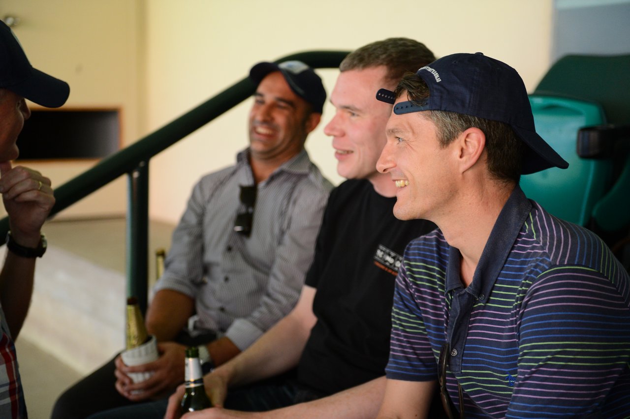 A group of men smiling and chatting while watching a cricket match, some holding drinks.
