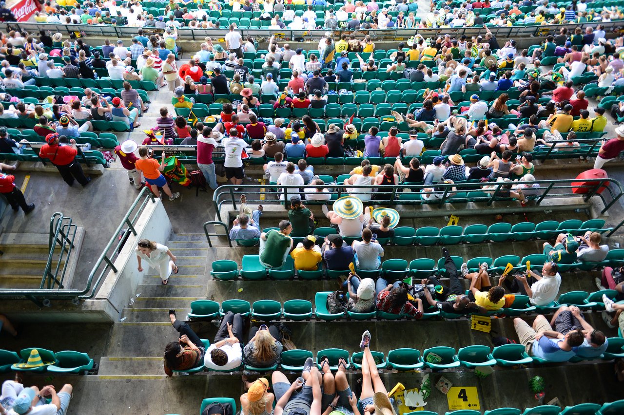 A large crowd of cricket fans sits in green stadium seats, wearing colorful hats and cheering during a match.