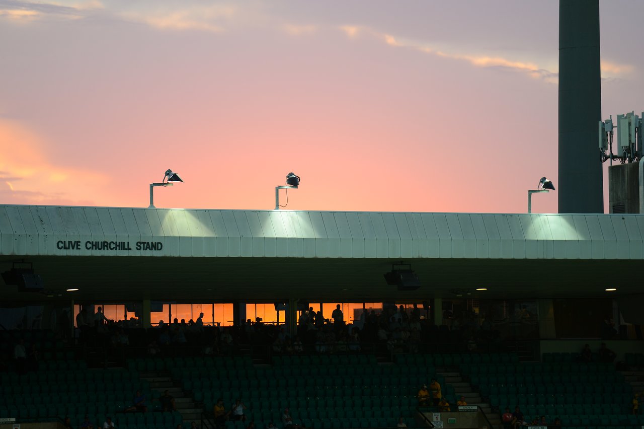 A cricket stadium stand with spectators, lit by sunset, with floodlights and birds perched on lamps.
