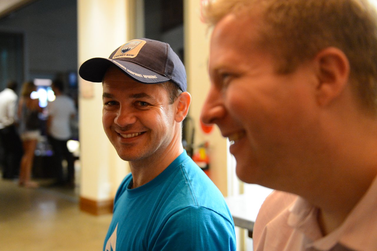 Two men smiling and talking indoors, one wearing a blue shirt and a cricket-themed cap.