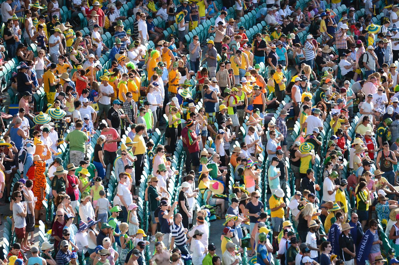 A large crowd of cricket fans, mostly in yellow and green, cheering and standing in stadium seats.