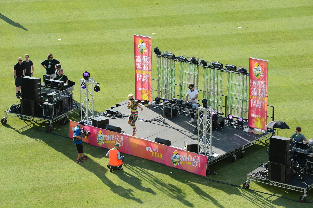 A DJ performs on an outdoor stage while a colorful performer engages with the audience at a cricket event.