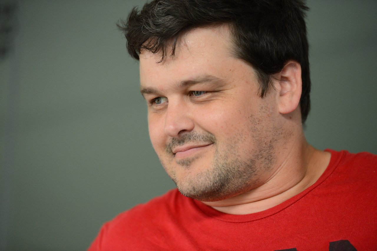 A man in a red shirt looks to the side with a slight smile.