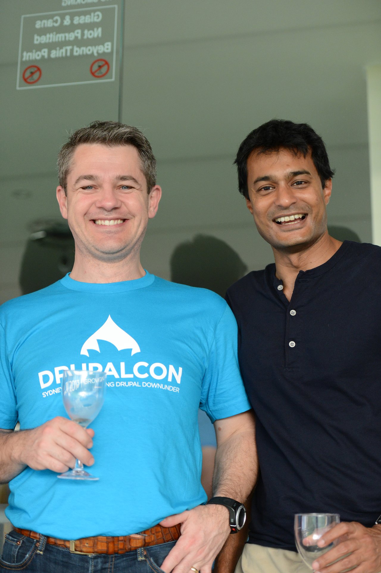 Two smiling men stand together, holding wine glasses, at DrupalCon Sydney 2013.