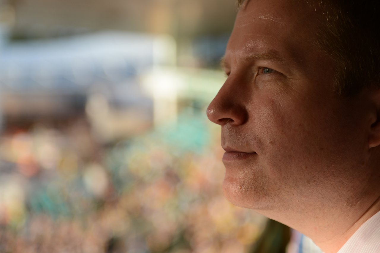 A man watches a cricket match with a focused expression, while the blurred crowd fills the background.