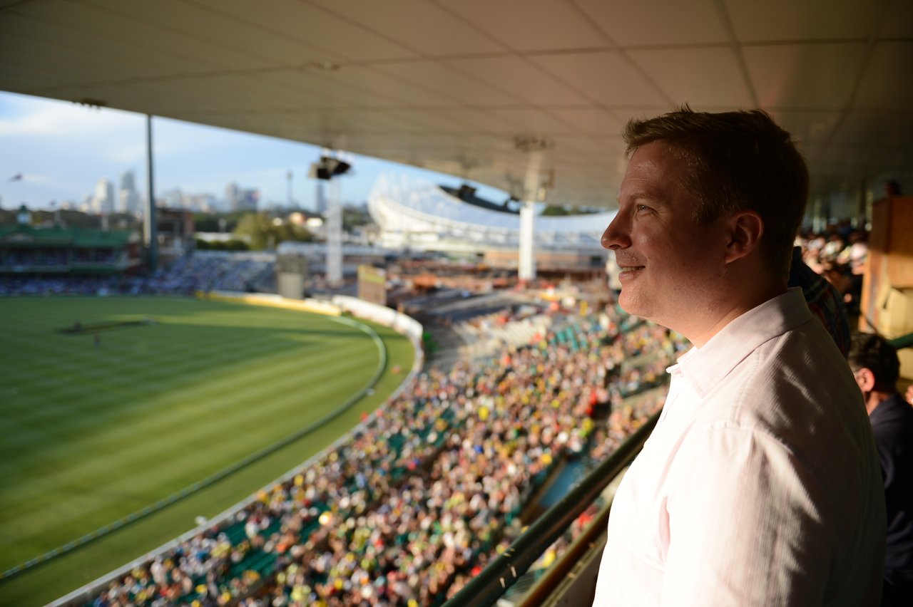 A man in a white shirt smiles while watching a cricket match from a stadium balcony.