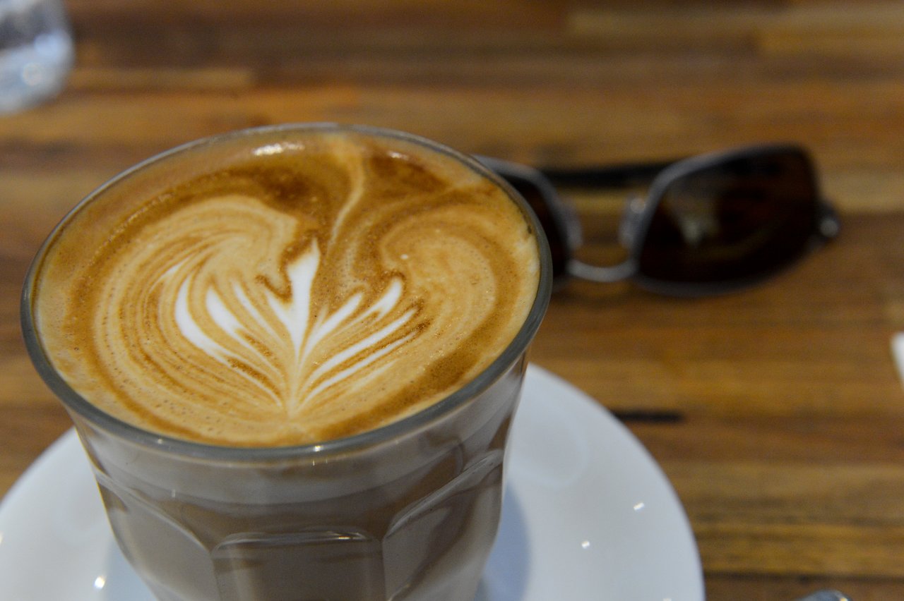 A glass of latte with latte art on a white saucer, placed on a wooden table with sunglasses nearby.