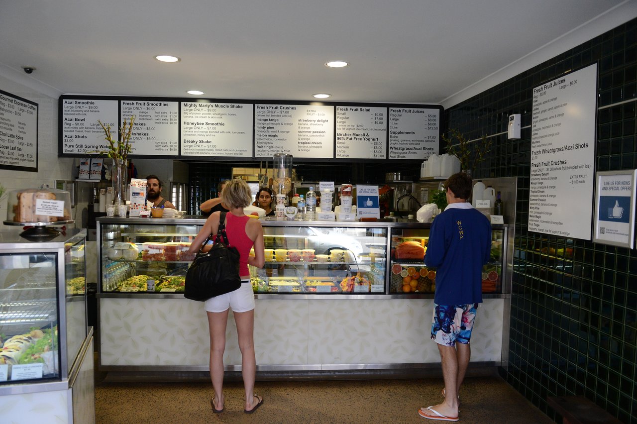 Two customers stand at a juice bar counter, ordering from staff behind the glass display filled with fresh ingredients.