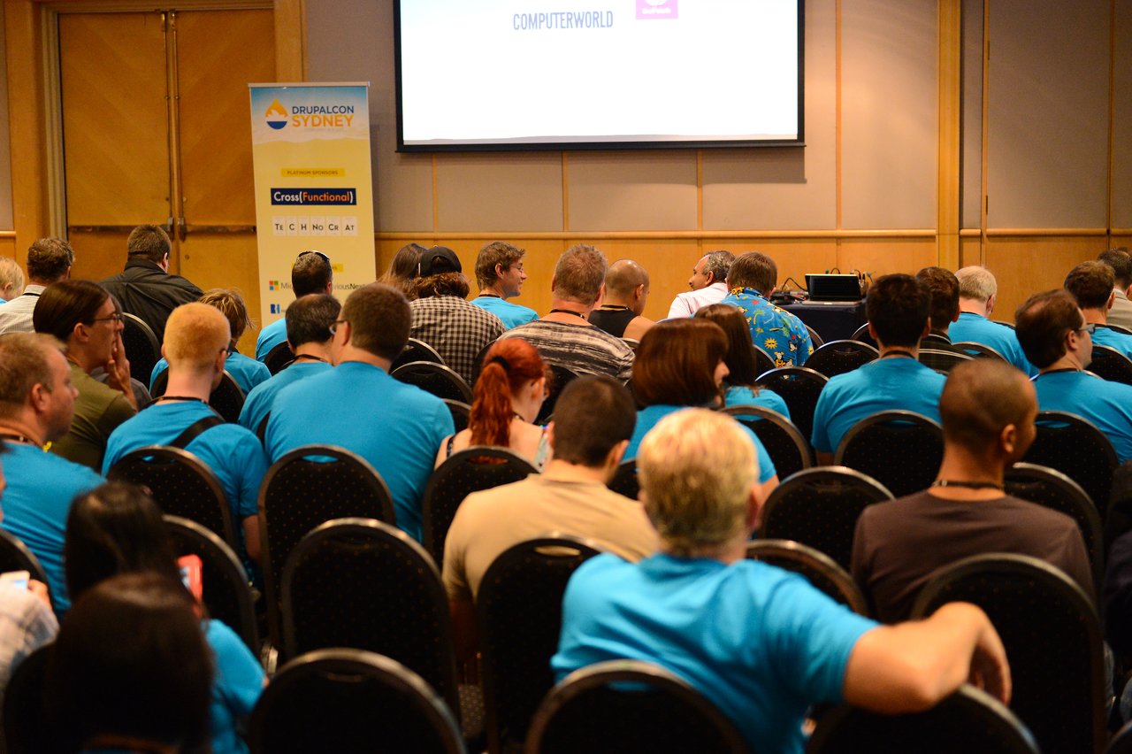 Audience members in blue shirts sit in rows at a DrupalCon Sydney 2013 conference session, facing the front.