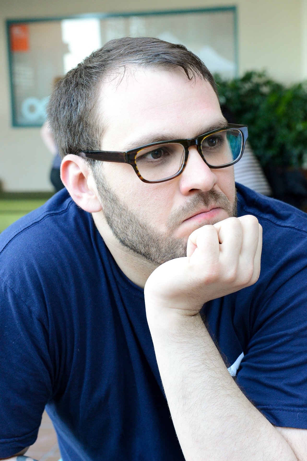 A man wearing glasses and a navy blue shirt rests his chin on his hand, looking thoughtful.