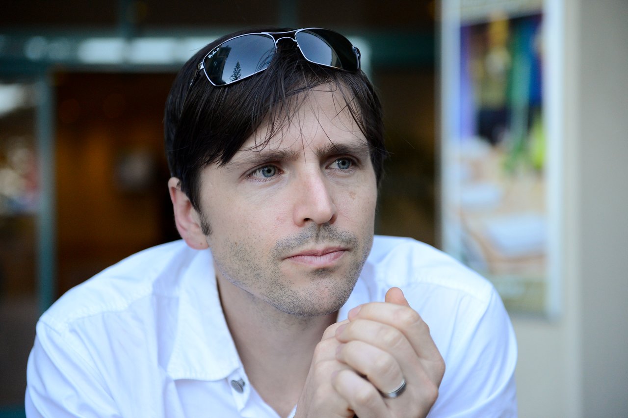A man in a white shirt with sunglasses on his head looks thoughtful while sitting at DrupalCon Sydney 2013.