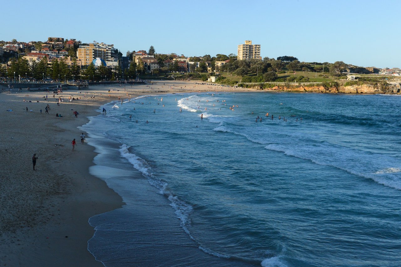 People are swimming and walking along a sandy beach with ocean waves and buildings in the background.