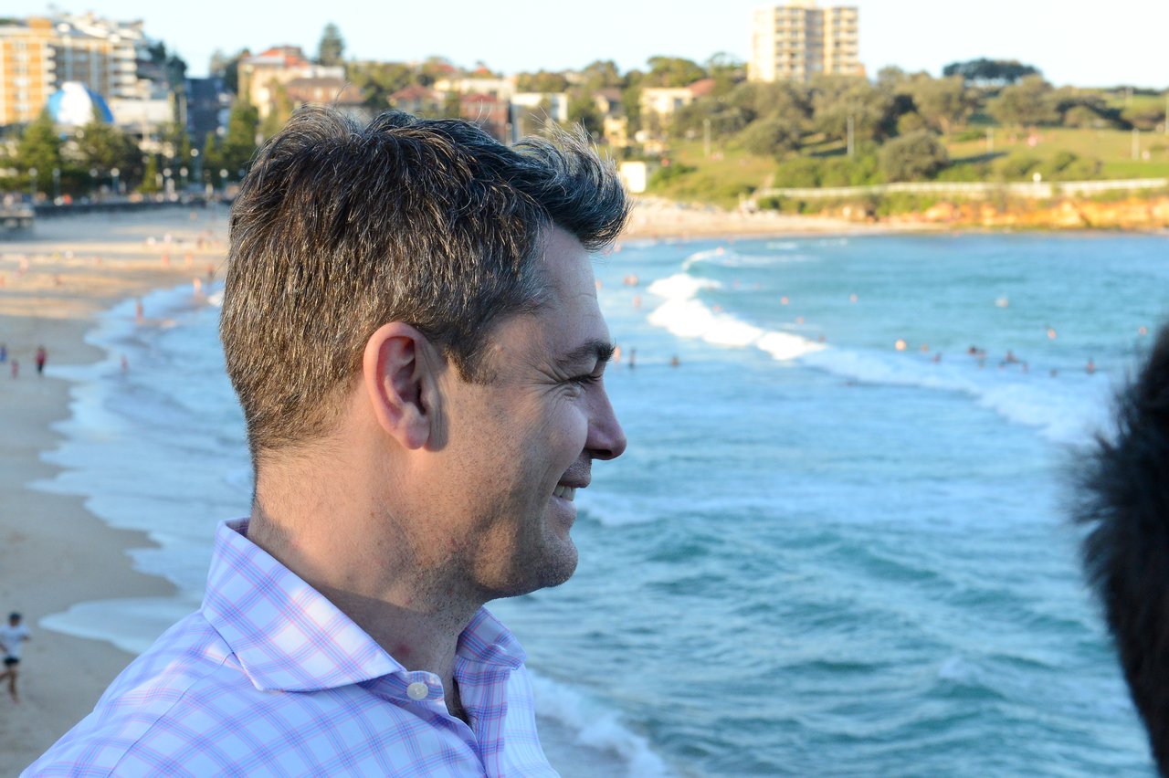 A man in a checkered shirt smiles while looking at the ocean during DrupalCon Sydney 2013.